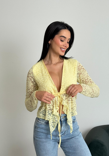 Mujer sonriendo usando un top de encaje amarillo y jeans, posando frente a una pared blanca.