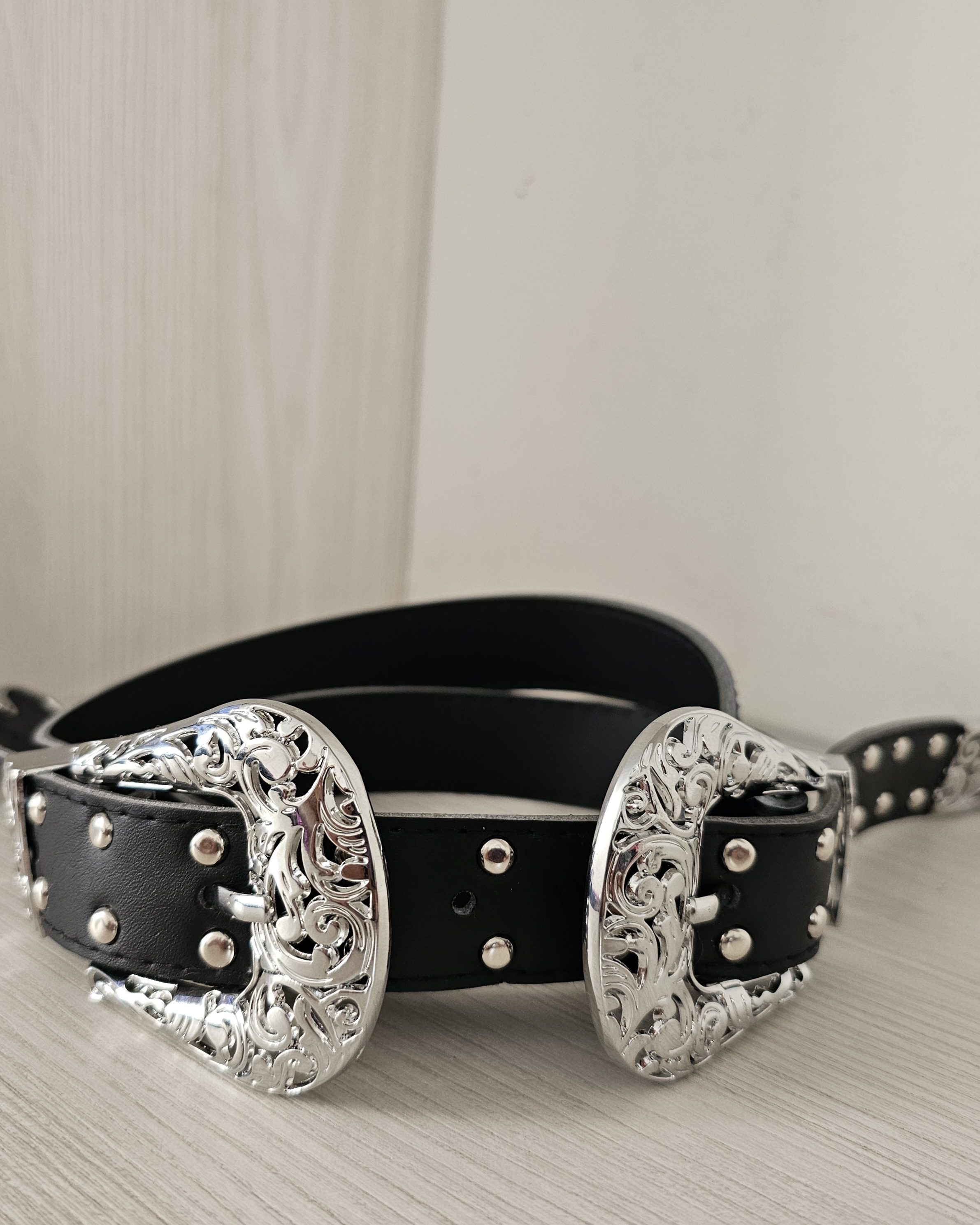 Cinturón negro para mujer de doble hebilla plateada con grabados vintage. Cinturón de cuero sintético con taches metálicos y punta decorativa labrada. Accesorio estilo western para jeans y pantalones sastre. Jessi Boutique.
